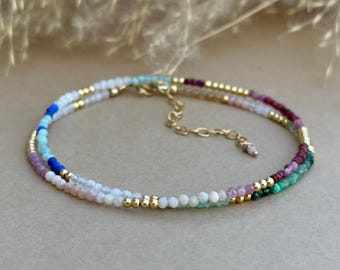 May include: A delicate multi-strand bracelet featuring small, round, multicolored gemstone beads in shades of white, blue, pink, green, and deep red, interspersed with tiny gold-toned metallic beads and a gold-toned chain extender with a clasp.