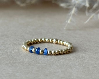 May include: A gold beaded ring with a row of small, vibrant blue gemstones. The ring is set against a neutral background, highlighting the contrast between the gold and blue. The ring is a delicate piece of jewelry.