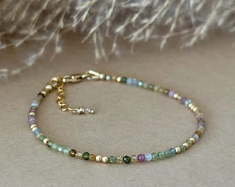 May include: Delicate gold bracelet featuring small, colorful beads in shades of green, purple, and blue. The bracelet has a gold clasp and is displayed on a neutral background. A stylish piece of jewelry.