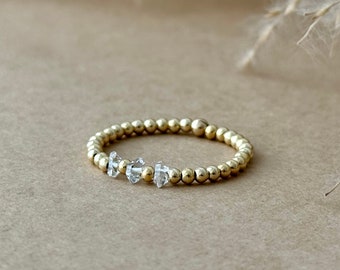 May include: A gold beaded ring with three clear, faceted gemstones. The ring is set against a neutral background with dried plants, highlighting the jewelry's delicate design and craftsmanship. The ring is a simple yet elegant piece.