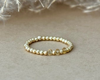 May include: A gold beaded ring with small, round beads and a few yellow-toned gemstones. The ring is set against a neutral background, highlighting its delicate design. This piece of jewelry is a simple yet elegant accessory.