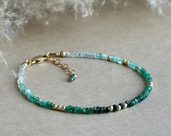May include: A delicate bracelet featuring a gradient of green and blue gemstones, transitioning to gold beads and a gold clasp. The bracelet is made of small, round beads and is designed to be worn around the wrist.