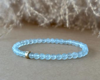 May include: A stretch bracelet composed of small, round, translucent light blue beads. The bracelet features a single, smooth, cylindrical gold-toned metal bead accent positioned asymmetrically along the strand of blue beads.