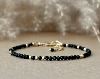 May include: Delicate black beaded bracelet with gold accents. The bracelet features small, faceted black beads interspersed with gold-colored beads and a gold clasp. The jewelry is displayed on a neutral background, highlighting the contrast of colors.