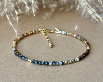 May include: Delicate beaded bracelet featuring small, multi-colored gemstones and gold-colored accents. The bracelet has a gold-colored clasp and is displayed on a neutral background. The gemstones include shades of blue, brown, and green.