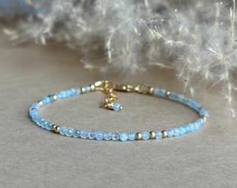 May include: Delicate bracelet featuring small, faceted, light blue beads interspersed with gold-colored beads. The bracelet has a gold-colored clasp and a small charm. The jewelry is set against a neutral background.
