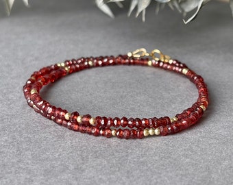 May include: A double-strand bracelet featuring small, faceted, deep red gemstones and interspersed gold-colored beads. The bracelet is secured with a gold-colored clasp. The jewelry is displayed on a gray surface, with a blurred background of white and green foliage.