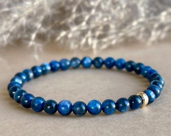 May include: A beaded bracelet featuring small, round, deep blue beads. A single gold-colored bead is incorporated into the design. The bracelet is displayed on a neutral-toned surface, with soft, blurred foliage in the background.