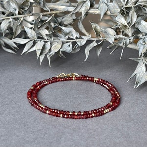 Op de afbeelding: Een delicate ketting met kleine, gefacetteerde, dieprode edelstenen en goudkleurige kralen. De ketting is gelaagd en vastgezet met een goudkleurige sluiting. De sieraden worden getoond op een grijze ondergrond met een vage, zilverkleurige botanische element op de achtergrond.