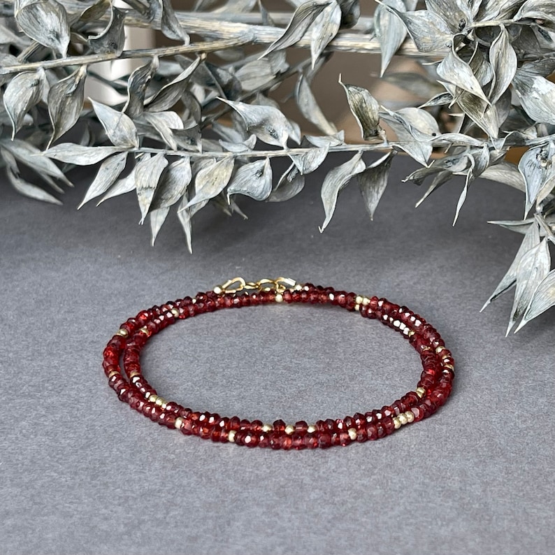 Op de afbeelding: Een delicate ketting met kleine, gefacetteerde, dieprode edelstenen en goudkleurige kralen. De ketting is gelaagd en vastgezet met een goudkleurige sluiting. De sieraden worden getoond op een grijze ondergrond met een vage, zilverkleurige botanische element op de achtergrond.