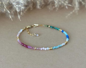 May include: A delicate bracelet featuring a gradient of small, faceted gemstone beads in shades of pink, purple, light blue, and deep teal, interspersed with tiny gold-toned metal beads. The bracelet includes a gold-toned metal lobster clasp and chain.