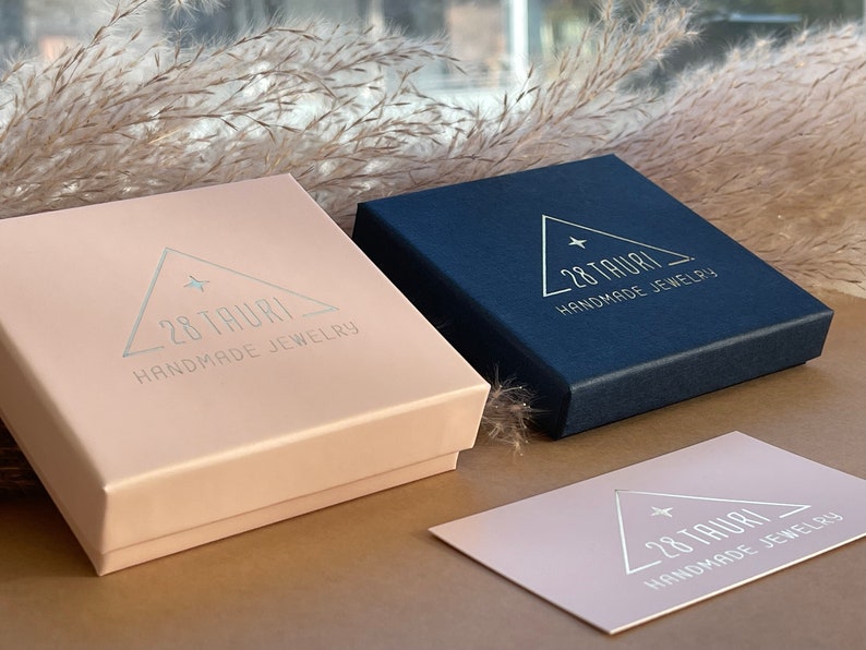 Op de afbeelding: Twee sieradendoosjes, &eacute;&eacute;n zachtroze en &eacute;&eacute;n marineblauw, met de tekst "28 Tauri Handmade Jewelry" op de deksels. Een bijpassende visitekaartje is ook zichtbaar. De dozen zijn rechthoekig en hebben een matte afwerking.