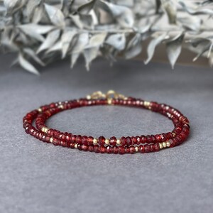 Op de afbeelding: Een gelaagde armband met kleine, gefacetteerde, dieprode edelstenen en goudkleurige kralen. De armband wordt getoond op een grijze ondergrond, met een vage achtergrond van gedroogd blad.