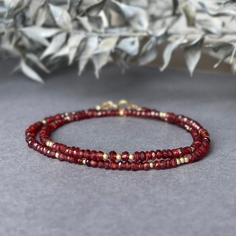Op de afbeelding: Een gelaagde armband met kleine, gefacetteerde, dieprode edelstenen en goudkleurige kralen. De armband wordt getoond op een grijze ondergrond, met een vage achtergrond van gedroogd blad.