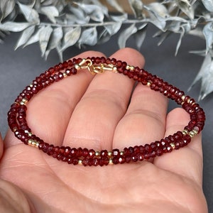 Op de afbeelding: Een dubbelstrengs armband met kleine, gefacetteerde, dieprode edelstenen en goudkleurige kralen. De armband is vastgezet met een goudkleurige sluiting. De sieraden worden gepresenteerd tegen een neutrale achtergrond.