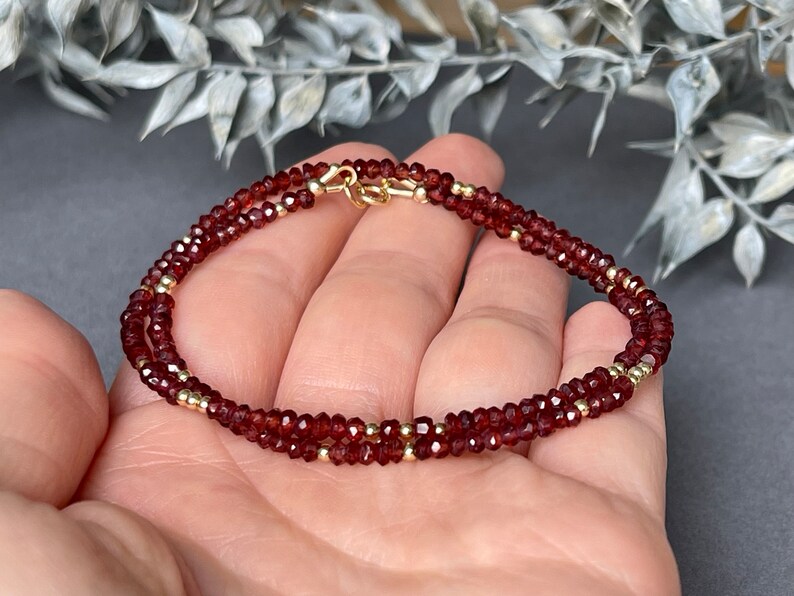 Op de afbeelding: Een dubbelstrengs armband met kleine, gefacetteerde, dieprode edelstenen en goudkleurige kralen. De armband is vastgezet met een goudkleurige sluiting. De sieraden worden gepresenteerd tegen een neutrale achtergrond.