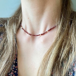 Op de afbeelding: Een delicate choker-ketting met kleine, dieprode kralen afgewisseld met kleine goudkleurige kralen. De ketting wordt getoond tegen een achtergrond van huid en vloeiend blond haar, wat de subtiele elegantie en het ontwerp benadrukt.