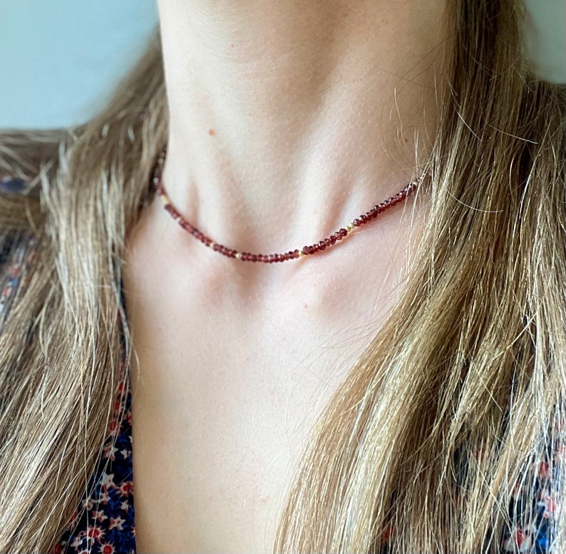 Op de afbeelding: Een delicate choker-ketting met kleine, dieprode kralen afgewisseld met kleine goudkleurige kralen. De ketting wordt getoond tegen een achtergrond van huid en vloeiend blond haar, wat de subtiele elegantie en het ontwerp benadrukt.