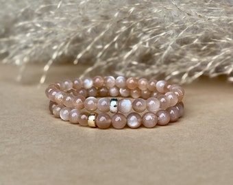 May include: Two stacked bracelets made of peach-colored, round gemstone beads. One bracelet has a silver accent bead, and the other has a gold accent bead. The background is a soft beige with feathery, light-colored accents.