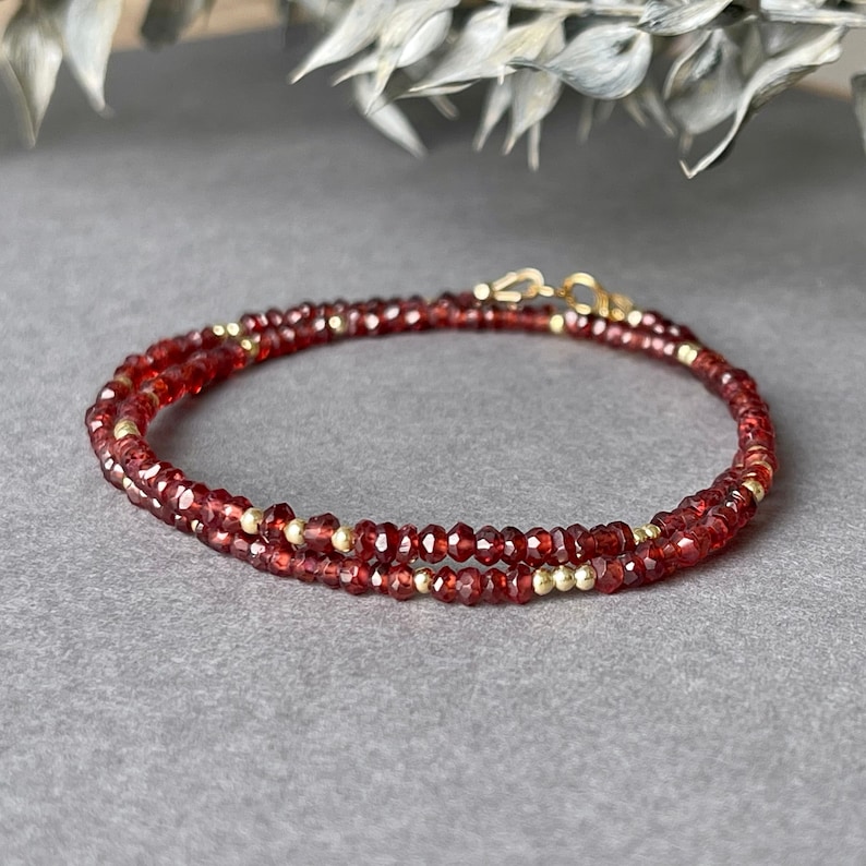 Op de afbeelding: Een dubbelstrengs armband met kleine, gefacetteerde, dieprode edelstenen en goudkleurige kralen. De armband is voorzien van een goudkleurige sluiting. De sieraden worden gepresenteerd op een grijze ondergrond, met een vage achtergrond van witte en groene bladeren.