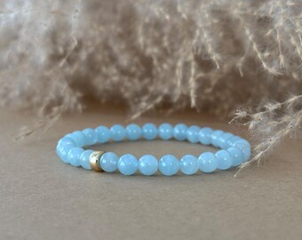 May include: A stretch bracelet composed of smooth, round, light blue beads. The bracelet features a single, small, polished gold-toned metal bead spacer positioned among the light blue beads, creating a subtle contrast in the design.