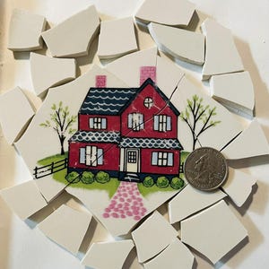 May include: A broken ceramic tile mosaic featuring a red house with a black roof, white windows, and a pink chimney. The house is surrounded by white broken pieces and a quarter coin for scale. The mosaic includes trees and a pink walkway.