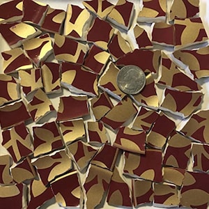 Könnte beinhalten: Eine Sammlung zerbrochener Keramikfliesen mit einem bordeauxroten und goldfarbenen Blattdesign. Die Fliesen sind unregelmäßig geformt und verstreut, mit einer Münze zum Größenvergleich. Das Goldblattmuster ist abstrakt.
