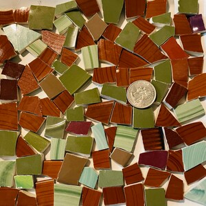 May include: A collection of irregularly shaped ceramic tiles in shades of brown, green, and white. A US quarter coin is included for size reference. The tiles have a variety of textures and patterns, suitable for mosaic art or craft projects.