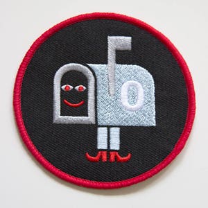 May include: An embroidered patch featuring a smiling mailbox with red shoes. The mailbox is white with a black face and is set against a black background with a red border.