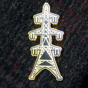 May include: A gold-coloured metal pin shaped like an electrical tower. The tower is outlined in black and has a black triangle in the centre.