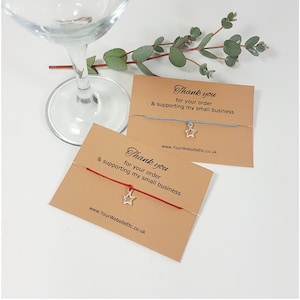 May include: Two star charm bracelets, one with a red cord and the other with a light blue cord, are presented on brown cards. The cards feature the text "Thank you for your order & supporting my small business" and a website address.