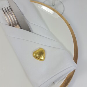 May include: A folded white linen napkin with a gold-colored heart-shaped accent on a white plate with a gold rim. A fork and knife are tucked into the napkin. The setting suggests a formal dining occasion.