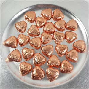 May include: A collection of heart-shaped candies wrapped in shiny copper-colored foil, arranged on a silver plate. The foil has a textured appearance, and the candies are evenly spaced. The image is well-lit, highlighting the metallic sheen of the foil.