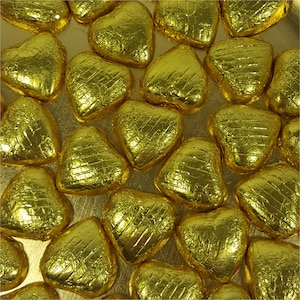 May include: Overhead shot of numerous heart-shaped candies wrapped in shiny gold foil. The foil has a textured appearance, and the candies are arranged closely together, creating a pattern across the surface.