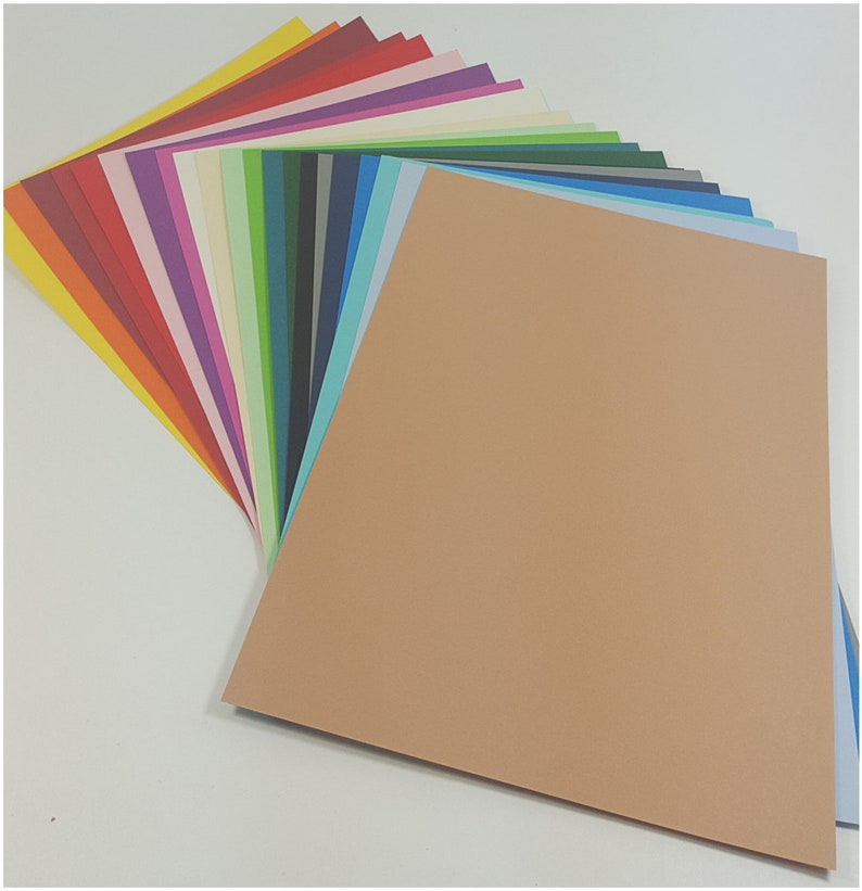 May include: A selection of coloured cardstock paper fanned out to display a range of hues. The colours include red, orange, yellow, green, blue, and purple. A large tan sheet of paper is in the foreground.