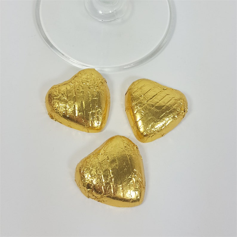 May include: Three heart-shaped chocolates wrapped in shiny gold foil. The chocolates are arranged on a white surface, with a portion of a clear glass visible at the top. The foil has a textured appearance.