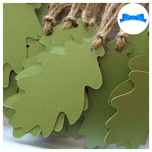 May include: A close-up shot of several green oak leaf-shaped tags with natural twine string. The tags are a muted green colour and have a textured surface. The twine is knotted at the top of each tag, suggesting they are ready for use as gift tags or decorative elements.