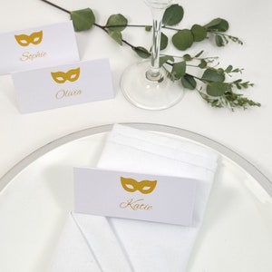 May include: White place cards with gold mask designs and names: Sophie, Olivia, and Katie. The cards are on a white plate with a silver rim, a white napkin, and greenery. A wine glass is also present.