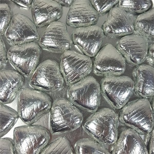May include: An overhead shot of numerous silver foil-wrapped heart-shaped candies. The candies are arranged closely together, filling the frame. The foil has a crinkled texture, reflecting light and adding visual interest. The overall theme is festive and celebratory.