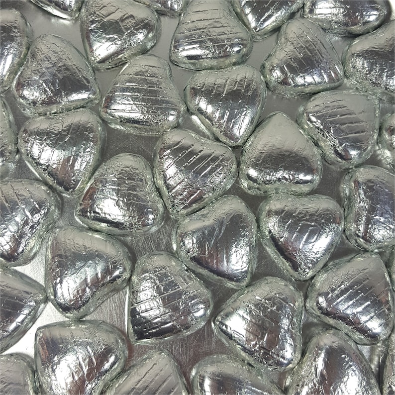 May include: An overhead shot of numerous silver foil-wrapped heart-shaped candies. The candies are arranged closely together, filling the frame. The foil has a crinkled texture, reflecting light and adding visual interest. The overall theme is festive and celebratory.