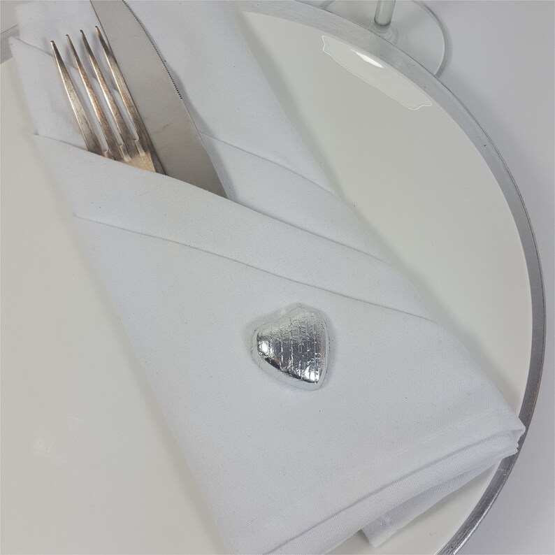 May include: A white folded napkin with a silver heart-shaped embellishment, a fork, and a knife on a white plate. The napkin is folded into a pocket shape, holding the silverware. The plate has a silver rim.