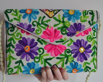 Può includere: Una pochette in tessuto bianco con un ricamo floreale. Il ricamo presenta fiori colorati in tonalità di rosa, viola, blu e verde. La pochette ha una chiusura a lembo e una tracolla a catena di perline.
