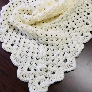 May include: A white crocheted shawl with a scalloped edge. The shawl is folded in half and laid flat on a wooden surface.