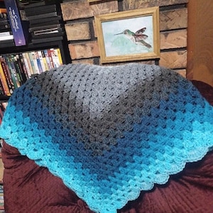 May include: A crocheted shawl in shades of blue and gray, arranged on a burgundy surface. The shawl features a scalloped edge and a gradient effect, transitioning from dark gray to light blue.