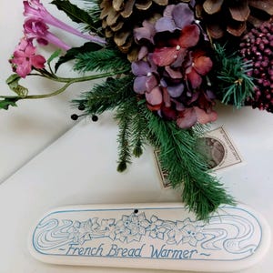 May include: A white ceramic French bread warmer with blue floral and wave designs. The text "French Bread Warmer" is written in blue. The warmer is decorated with a floral arrangement of purple and pink flowers, pinecones, and greenery.