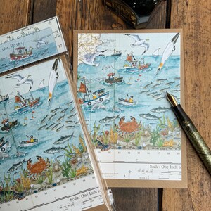 May include: Greeting cards with a nautical theme, featuring illustrations of fishing boats, seagulls, fish, and crabs in a marine environment. The cards are packaged with envelopes. A fountain pen rests on the surface.