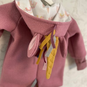 Puede incluir: Una sudadera con capucha rosa con un cuerno y una cola de unicornio hechos de fieltro. La capucha está forrada con una tela blanca con un patrón de arcoíris.