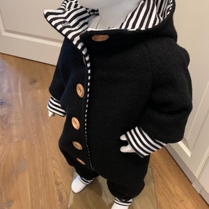 Puede incluir: Mono de bebé negro con capucha y botones de madera. El interior y el ribete presentan un estampado de rayas blancas y negras. Los puños y los tobillos también tienen el diseño de rayas. El mono se exhibe en un maniquí.
