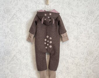 May include: A brown hooded baby onesie with a deer design. The onesie has a pink hood with two small ears and a tail. The onesie is decorated with white felt dots.
