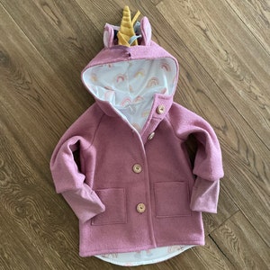 May include: A pink hooded jacket with a unicorn horn on the hood. The jacket has two pockets and a button closure. The lining of the hood is white with a rainbow pattern.